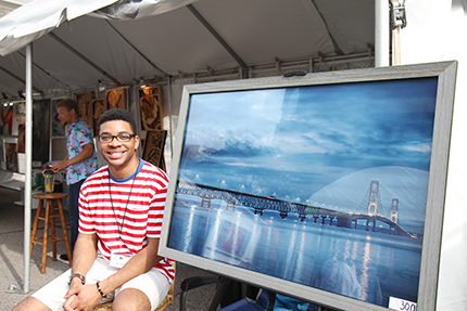 WCC student Spenser Williams sits near his work at the New Art, New Artist booth at the Ann Arbor Street Art Fair.