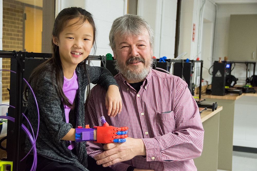 Washtenaw Community College faculty member Tom Penird with Lucy, an 8-year-old Ann Arbor girl whose prosthetic hand was created using 3D printers in WCC’s Industrial Technology laboratories. | Photo by CJ South